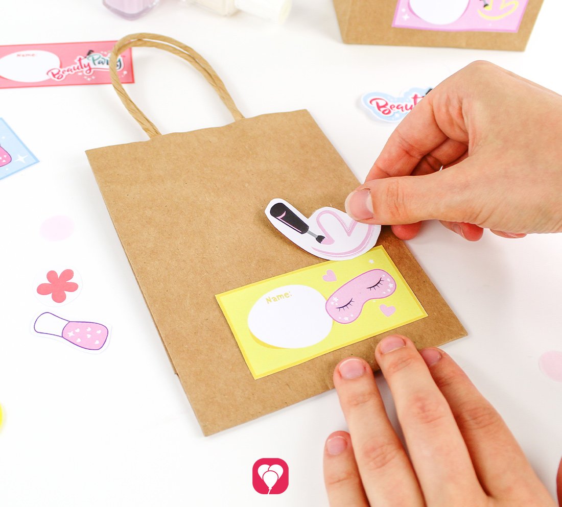 Es werden das Namensschild sowie einzelne bunte Sticker im Design der balloonas Beauty Party Geschenkaufkleber auf eine braune Geschenktüte mit Henkel geklebt.